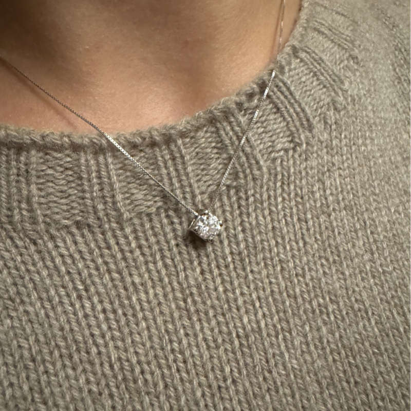 primo piano di un punto luce collana con diamante brillante montato su una sottile catenina d’oro bianco, indossata su un maglione a trama lavorata. 