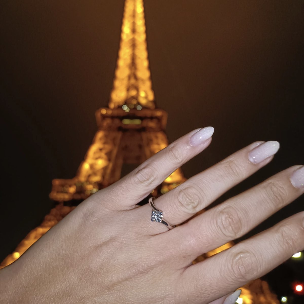 Una mano con un elegante anello solitario da 0.50 carati viene mostrata davanti alla Torre Eiffel illuminata di notte. L’immagine cattura il momento romantico di una proposta di matrimonio a Parigi, con lo sfondo sfocato che enfatizza il gioiello.